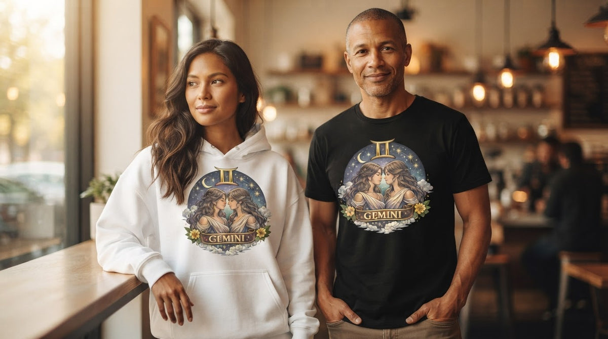 A couple in a cafe wearing the Gemini zodiac design, she in a white hoodie and he in a black tee, each showing two twins under a gold Gemini symbol, crescent moon and stars