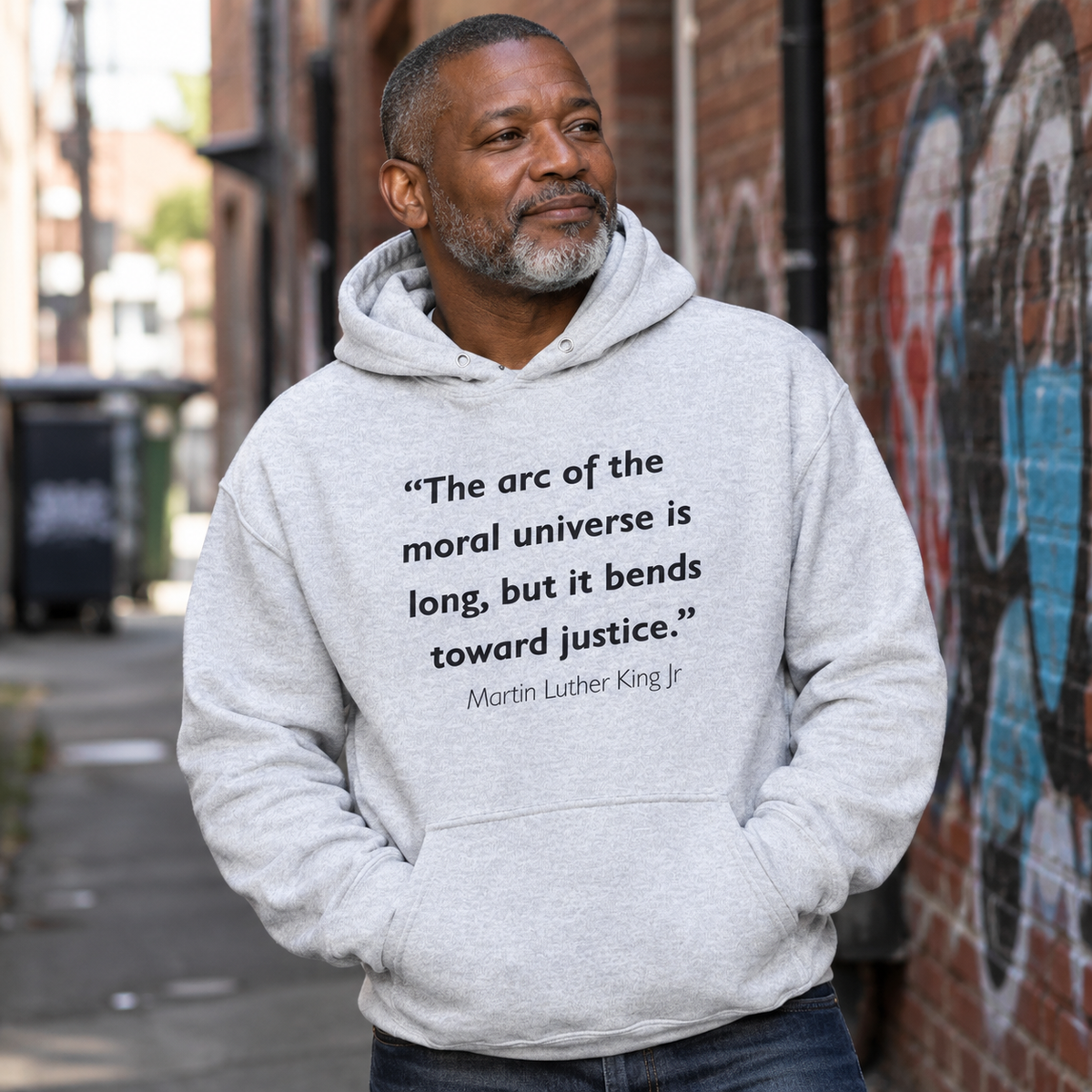 Man in a heather gray hoodie printed with the Martin Luther King Jr. quote "The arc of the moral universe is long, but it bends toward justice," standing in a city alley.